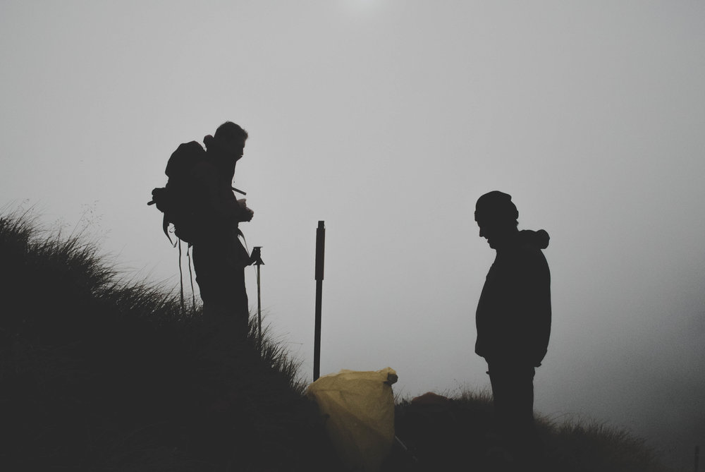 The silhouette of two backpackers on a hill with fog behind them.